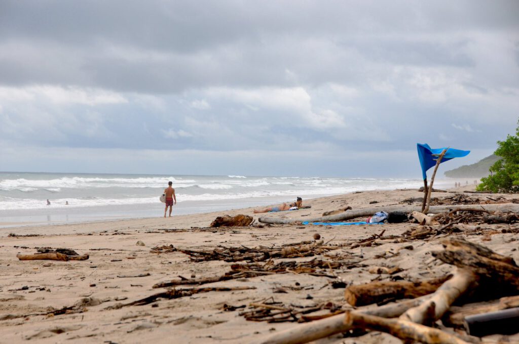 Weitläufiger, heller Sandstrand in Santa Teresa mit Treibholz und vereinzelten Surfern; im Hintergrund raue Wellen unter grauem Himmel. Am Strand weht die blaue Flagge, die international für herausragende Wasserqualität, Umweltmanagement und Sicherheit steht.