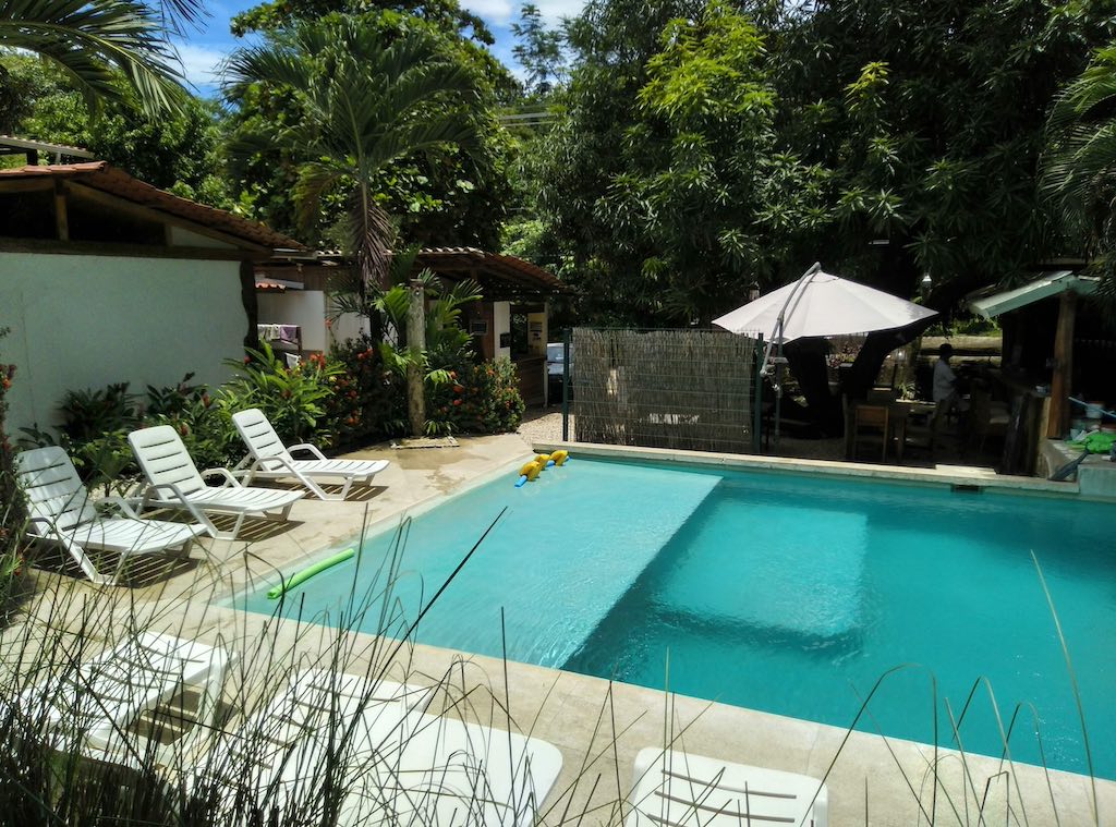 Ein Pool mit türkisgrünem Wasser, am Rand Liegen im schatten, ein Sonnenschirm und im Hintergrund Bäume