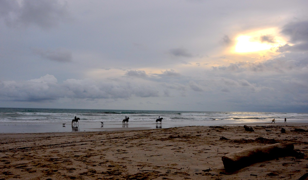 Strand von Santa Teresa bei bewölktem Himmel in der Abendsonne; drei Reiter auf Pferden bewegen sich entlang der Brandung im Gegenlicht.